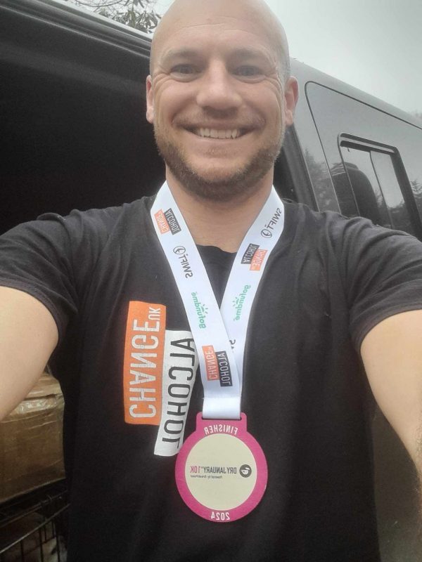Charity wooden medal for Alcohol Change UK 2024. Event orgainser wearing a bespoke wooden medal with big smile and printed lanyard showing sponsors logos including Swift Event Supplies.