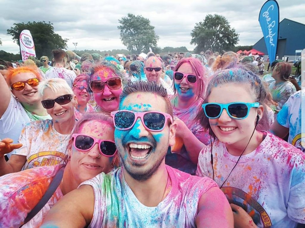 Team of runners wearing sunglasses at a colour run event for charity supporting Sue Ryder charity. Happy runnenrs wearing plain white t-shirt covered in holi colour powder.
